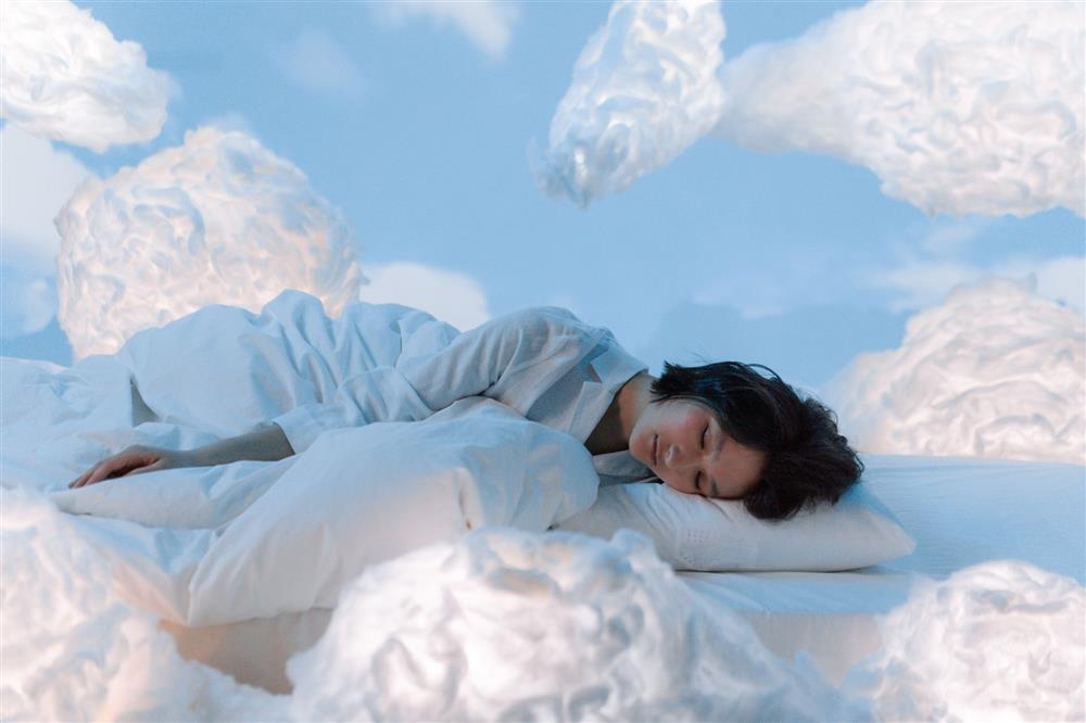 Person sleeping comfortably in a white bed surrounded by soft, cloud-like shapes in a calming blue bedroom.